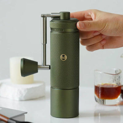 Green portable coffee grinder on a white surface with coffee beans and a glass of coffee.