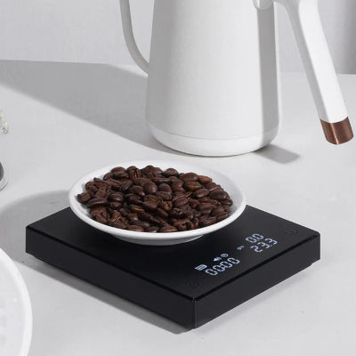 White ceramic kettle on a white surface with a bowl of coffee beans on a digital scale.