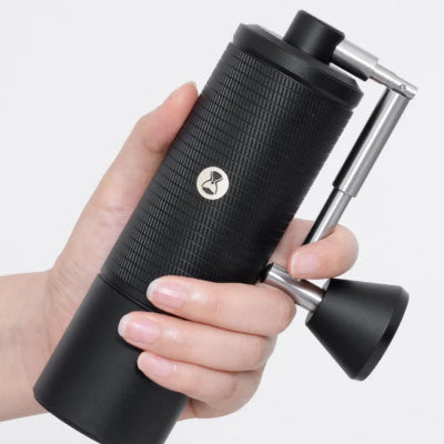 A hand holding a black manual coffee grinder with a textured grip and silver metal handle against a light gray background.