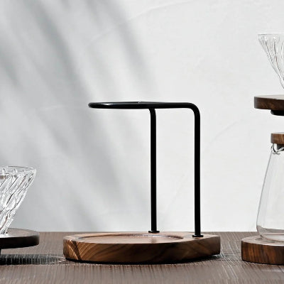 A minimalist black metal coffee pour-over stand with a round wooden base, styled on a wooden table with coffee glassware in soft shadowed light.