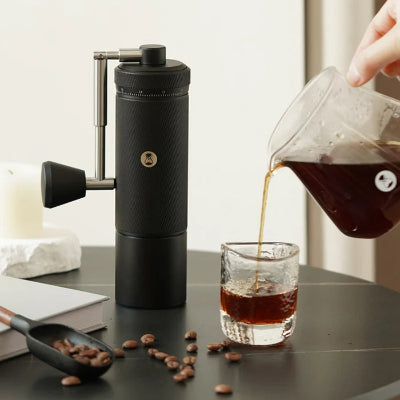 Hand pouring coffee from a glass carafe into a glass on a table with a coffee grinder and beans.