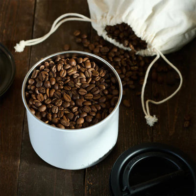 White container filled with coffee beans on a wooden surface with a drawstring bag of coffee beans.