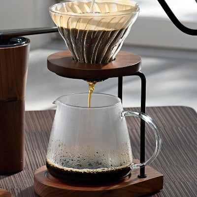 A pour-over coffee setup with a glass cone filter on a wooden dripper stand, with hot water being poured from a black gooseneck kettle into the filter above a glass carafe, set on a wooden table.