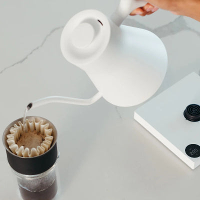 Person pouring coffee from a white pitcher into a glass container with a filter on a light gray surface.