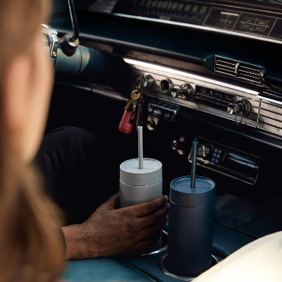 Person sitting in a vintage car with two blue cups on the dashboard.
