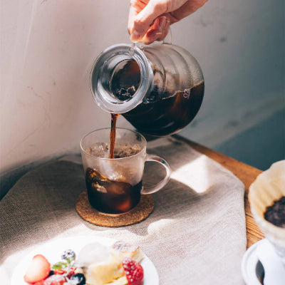 Person pouring coffee from a French press into a glass on a table with a plate of fruit.