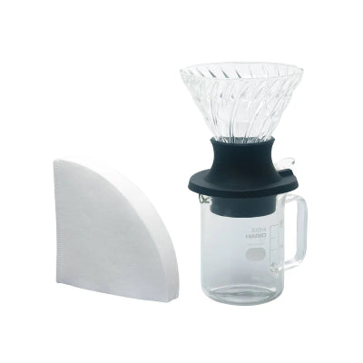A clear glass pour-over coffee dripper with a black collar, shown with a white paper filter on a plain white background.
