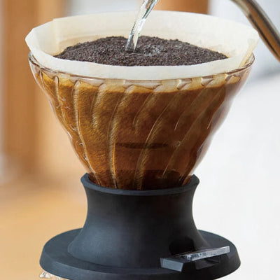 A close-up of a pour-over coffee maker with hot water being poured from a metal gooseneck kettle into a glass cone filter filled with coffee grounds, set on a black base.