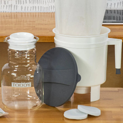 A coffee brewing set on a wooden table, including a clear glass carafe with a white lid and Toddy branding, a white plastic dripper and pot, a dark gray silicone sleeve, and two white filter discs.