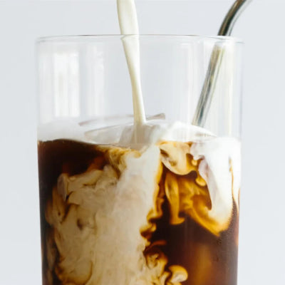 A clear glass tumbler filled with iced coffee and milk swirling inside, with a metal straw, on a light gray background.