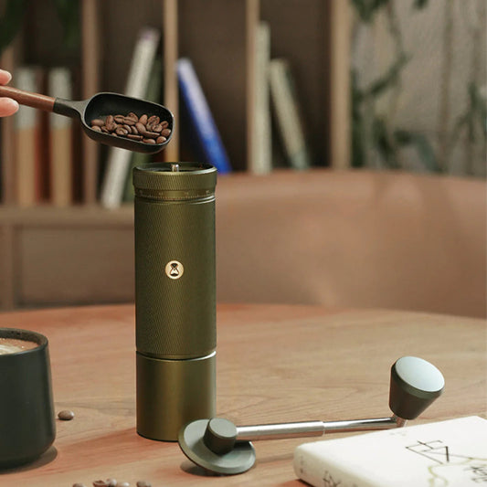 Green portable coffee grinder on a wooden table with coffee beans and a notebook.