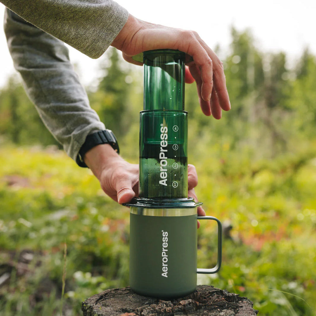 Person using an Aeropress coffee maker outdoors on a tree stump with a natural background.
