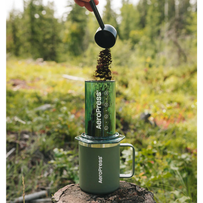 A person using an AeroPress outdoors on a stump with a forest background