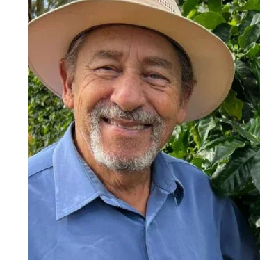 Man wearing a wide-brimmed hat and blue shirt standing in front of coffee plants with green beans.