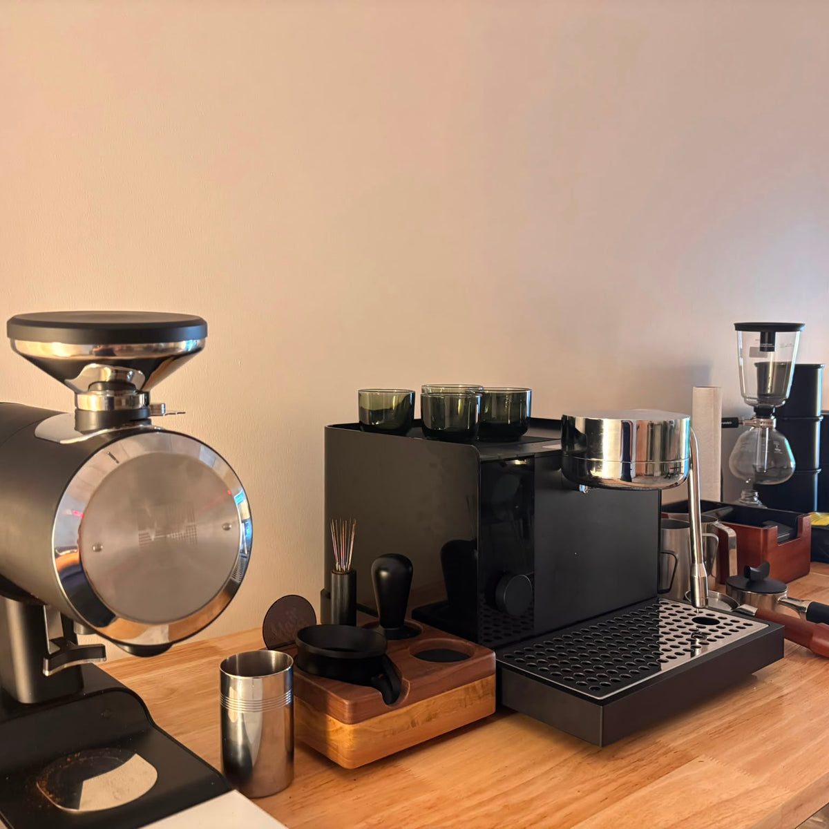 A countertop coffee station setup with a large black and silver coffee grinder in the foreground, a black espresso machine, a wooden cutting board with metal accessories, and additional coffee equipment in the background against a plain beige wall.