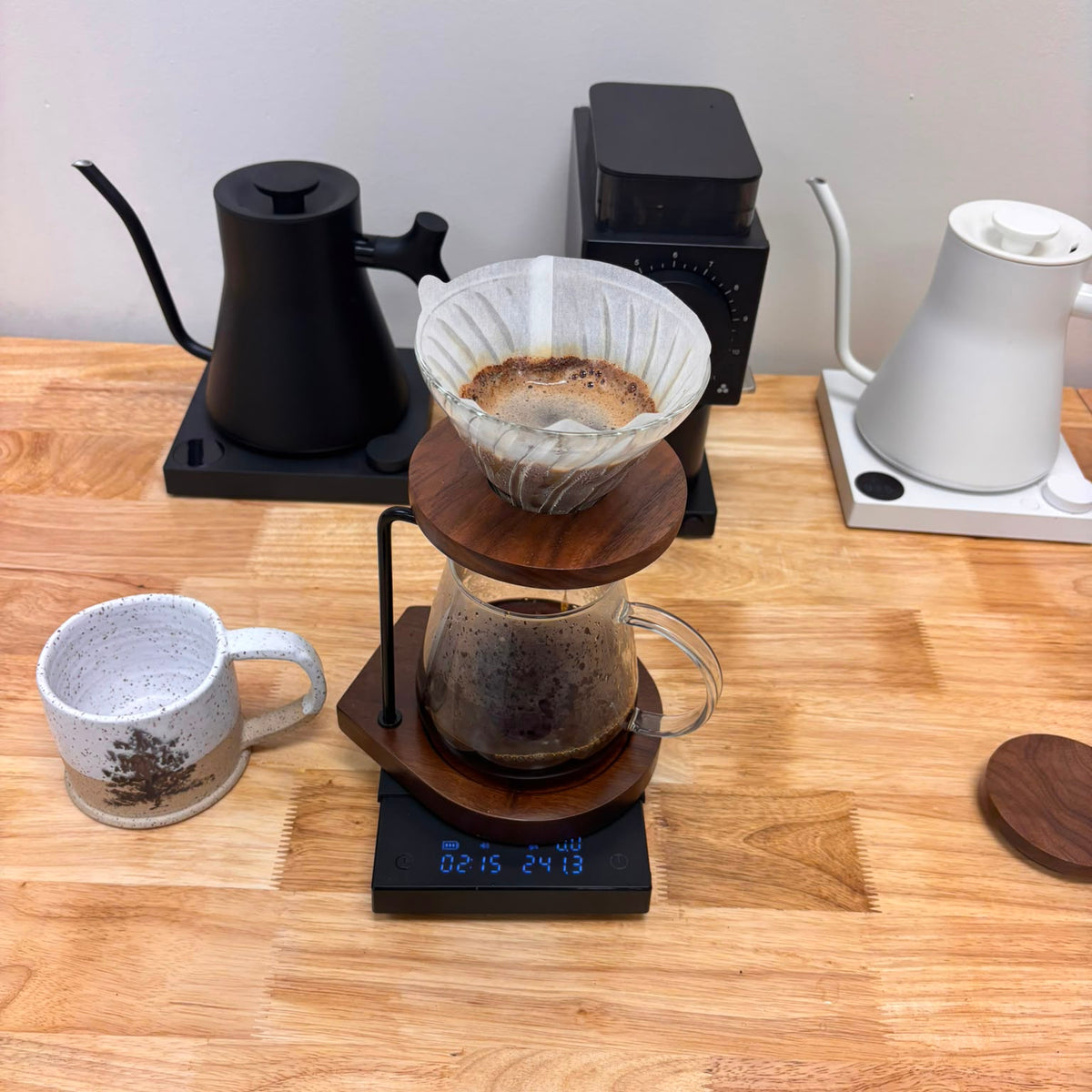 A pour-over coffee setup on a wooden table, featuring a glass carafe with dark coffee, a wooden percolator, a white dripper, a digital scale, a black go-kettle, and a white kettle on a white base.