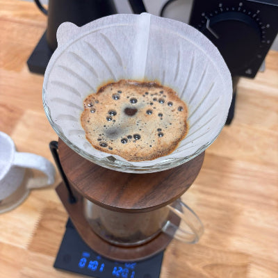A top-down view of a pour-over coffee setup on a wooden table, featuring a white paper filter cone over a glass carafe resting on a digital scale, with brewed coffee inside.