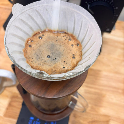 A close-up of a pour-over coffee setup with coffee dripping into a glass carafe on a wooden base, placed on a digital scale over a light wood tabletop.