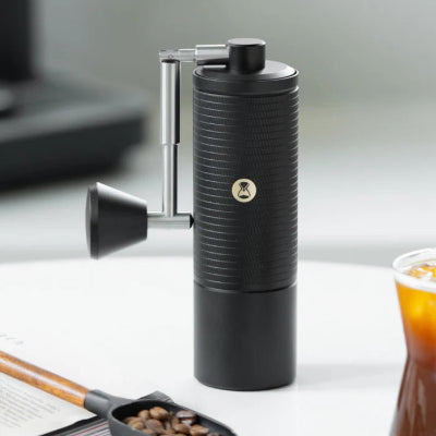 A black manual coffee grinder with a silver handle and a small gold logo, standing on a white tabletop next to coffee beans, a scoop, and a glass of iced coffee.