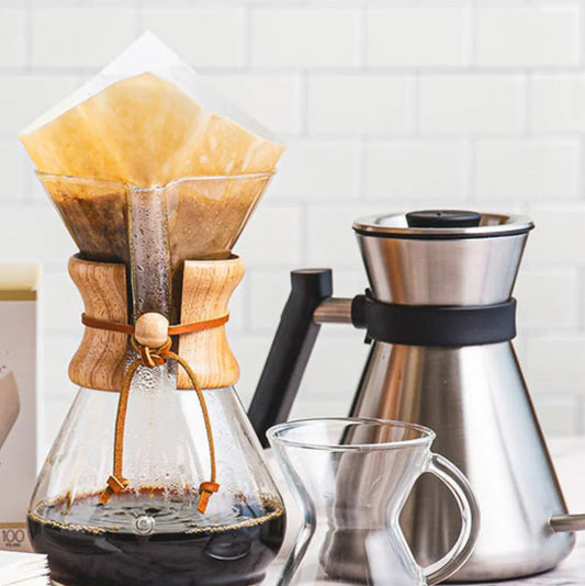 Coffee brewing setup with a wooden Chemex, stainless steel carafe, and glass mug on a marble surface.
