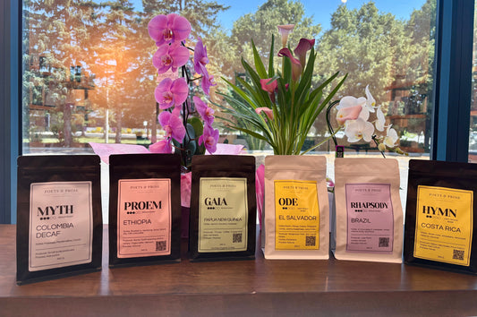 Six coffee bags from Poets & Prose Coffee Roasters lined up on a wooden shelf in front of a window with pink and white orchids in the background. The bags feature different label colors: black, pink, olive green, yellow, lavender, and black.