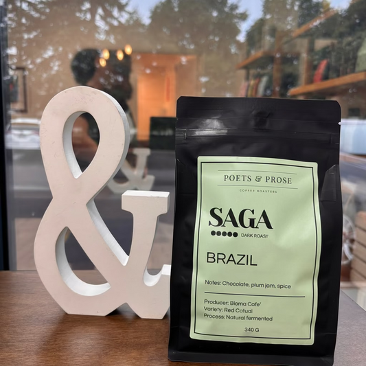 A black bag of Saga Coffee Brazil whole bean coffee stands on a wooden table next to a large white decorative ampersand. The bag features a pale green label with black text.