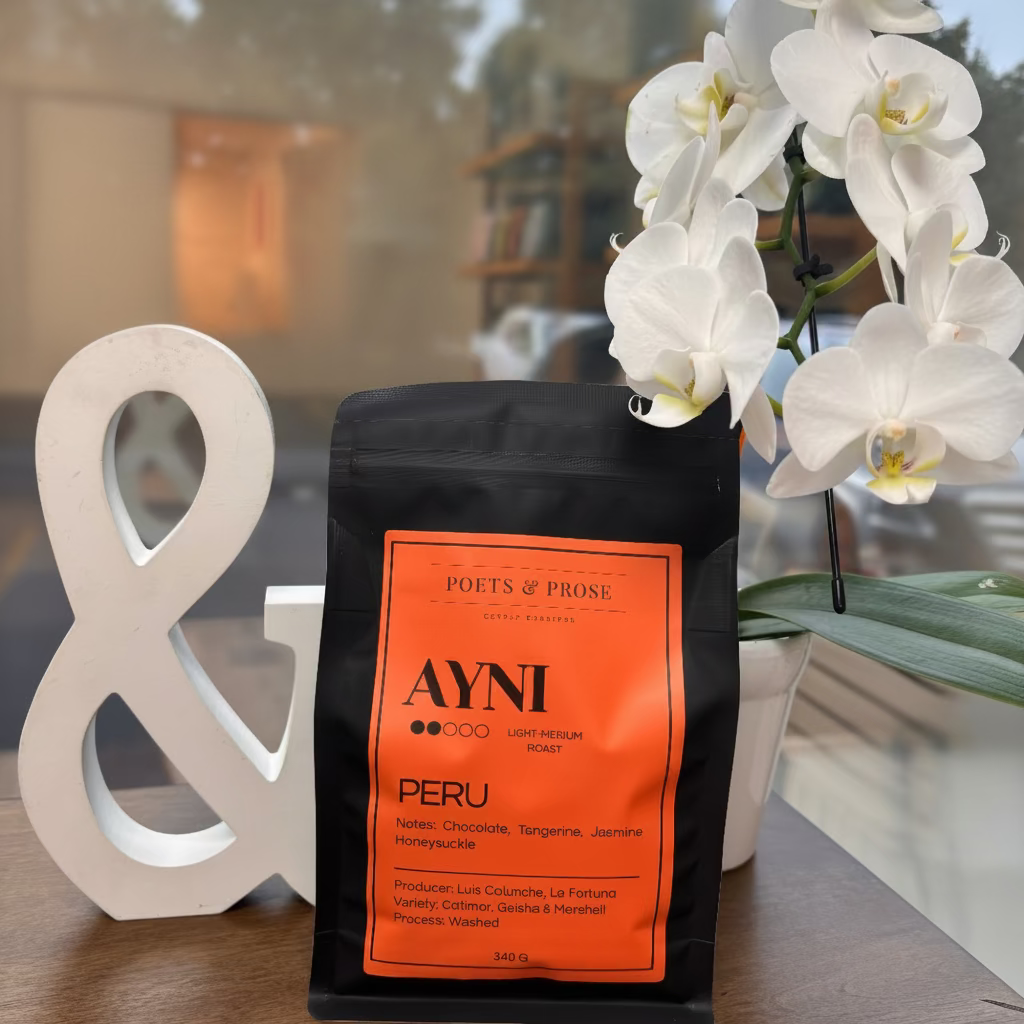 A black bag of Poets & Prose Coffee Roastery Peru coffee sits on a wooden table, styled with a white decorative ampersand and white orchids in front of a window.