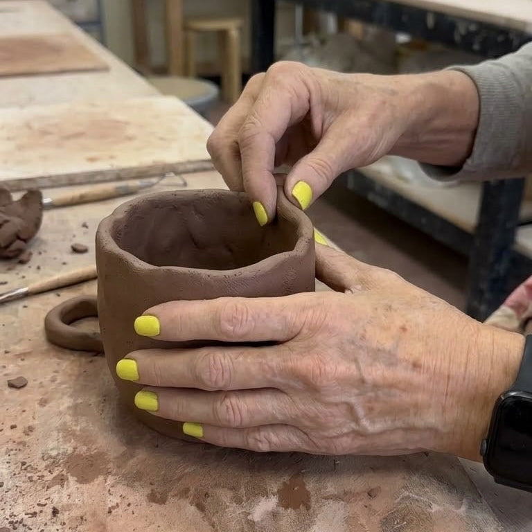 A close-up of a person's hands with bright yellow nail polish shaping a small brown clay mug on a pottery workbench.