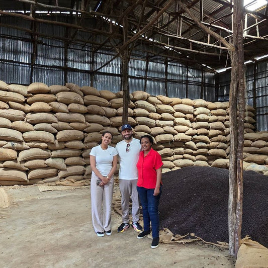 Three people standing in a large warehouse filled with stacked beige sacks and a pile of dark brown to black dried plant material, likely coffee beans or similar agricultural product.