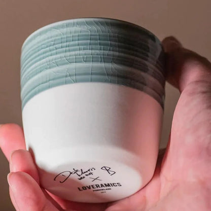 A hand holding a small ceramic cup or candle container with a textured sage green top half and a smooth white bottom half. The base is printed with the brand name 'LOVERAMICS' and a signature.