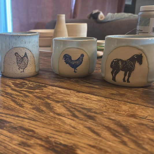 Five handmade ceramic mugs with rustic distressed finishes, each featuring a painted animal design (horse, chicken, rooster, horse, and horse), arranged on a wooden table.
