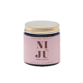 A small glass jar of roasted matcha with a light pink label and a gold lid, centered on a light beige circular background.