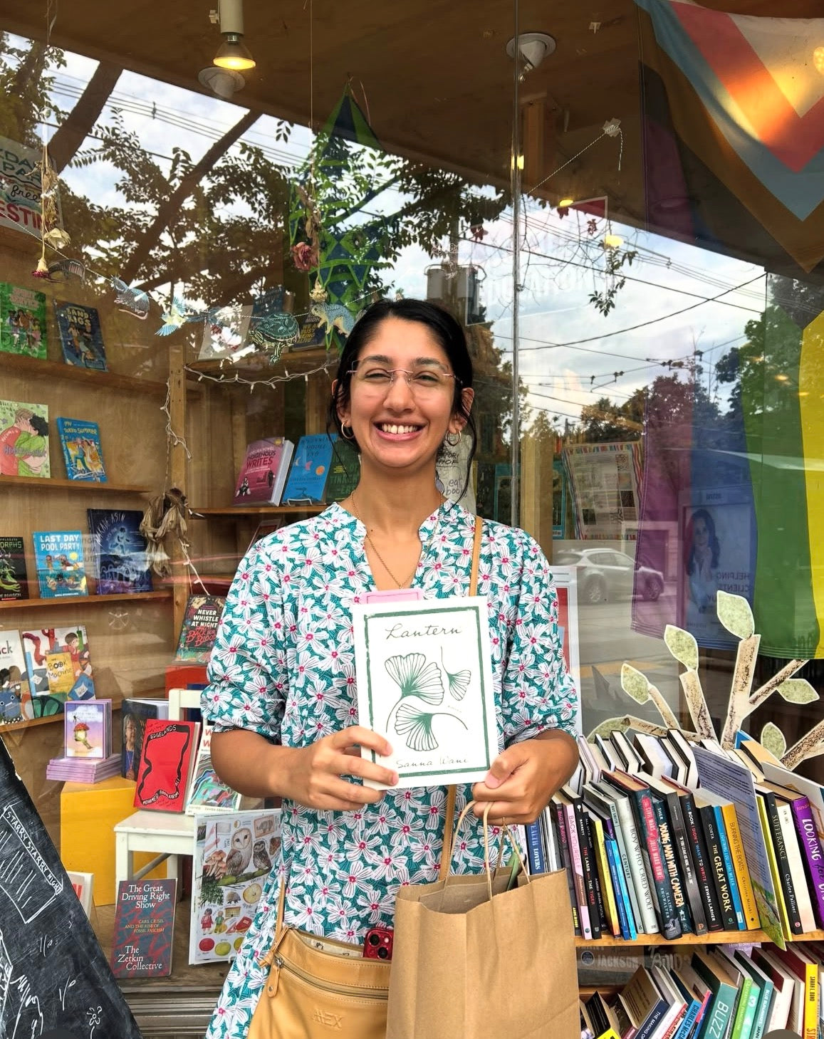 A smiling woman wearing glasses and a blue floral shirt stands in front of a bookshop window, holding a framed art print featuring a green ginkgo leaf with the word 'Lantern' written inside a white border.
