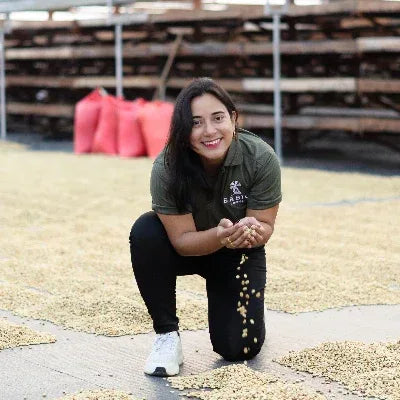 Woman kneeling on a surface with coffee beans, wearing a green shirt with text and black pants.