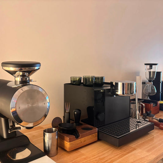 A countertop coffee station setup with a large black and silver coffee grinder in the foreground, a black espresso machine, a wooden cutting board with metal accessories, and additional coffee equipment in the background against a plain beige wall.