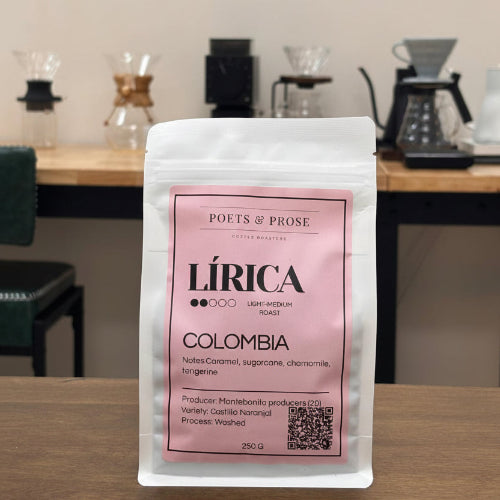 A 250g bag of 'Lírica' Colombia coffee from 'Poets & Prose' stands on a wooden table in a cafe setting with coffee makers blurred in the background.