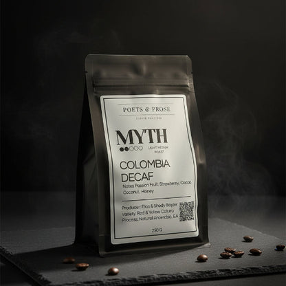 A black stand-up coffee bag labeled 'MYTH COLOMBIA DECAF' by Poets & Prose, placed on a wooden table with a blurred coffee-making setup in the background.