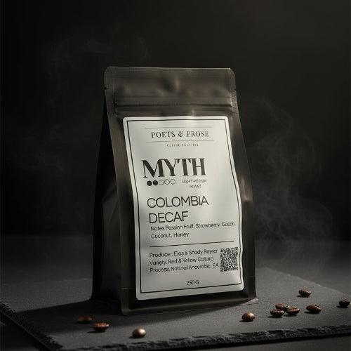 A black stand-up coffee bag labeled 'MYTH COLOMBIA DECAF' by Poets & Prose, placed on a wooden table with a blurred coffee-making setup in the background.