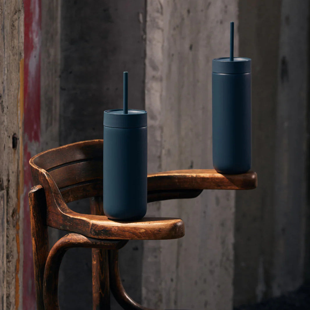 Two dark blue tumblers with straws on a rustic wooden chair against a textured wall.