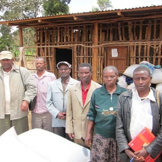 Group of people standing in front of a wooden building with bags of grain.