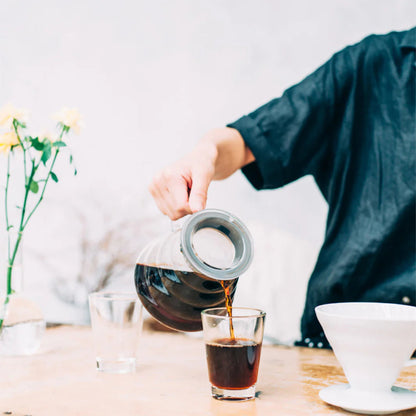 Person pouring tea from a glass teapot into a small glass on a wooden table.