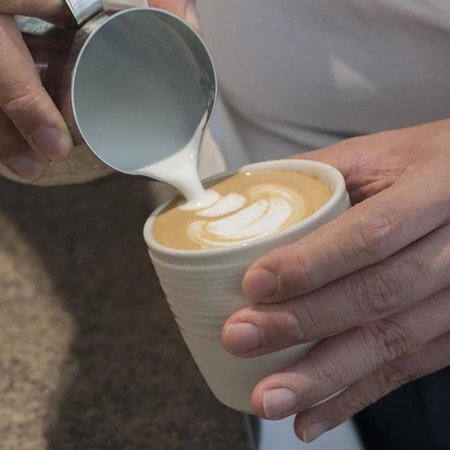 A close-up of a barista pouring steamed milk from a metal pitcher into a white ceramic cup of coffee with latte art, with the barista's hands visible.