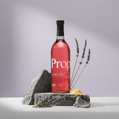 A bottle of Prop Lavender syrup stands on gray stones with dried lavender sprigs and a small pile of yellow powder against a soft lavender-gray studio background.