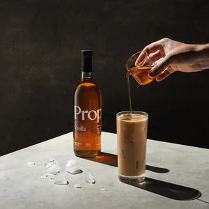 A hand pours amber liquor from a small glass into a tall glass of iced coffee, with a bottle of Proper syrup on a dark tabletop against a dark background.