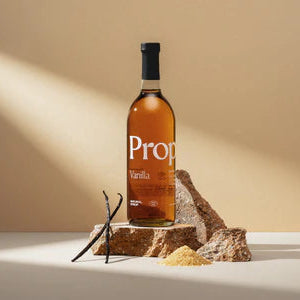 A bottle of Prop Vanilla syrup stands on a rustic stone slab with vanilla beans and a scoop of vanilla extract in front, set against a warm beige background with dramatic diagonal light rays.
