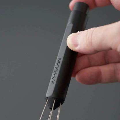 A close-up of a hand holding a black Anti-Minimal whisk against a dark gray background.