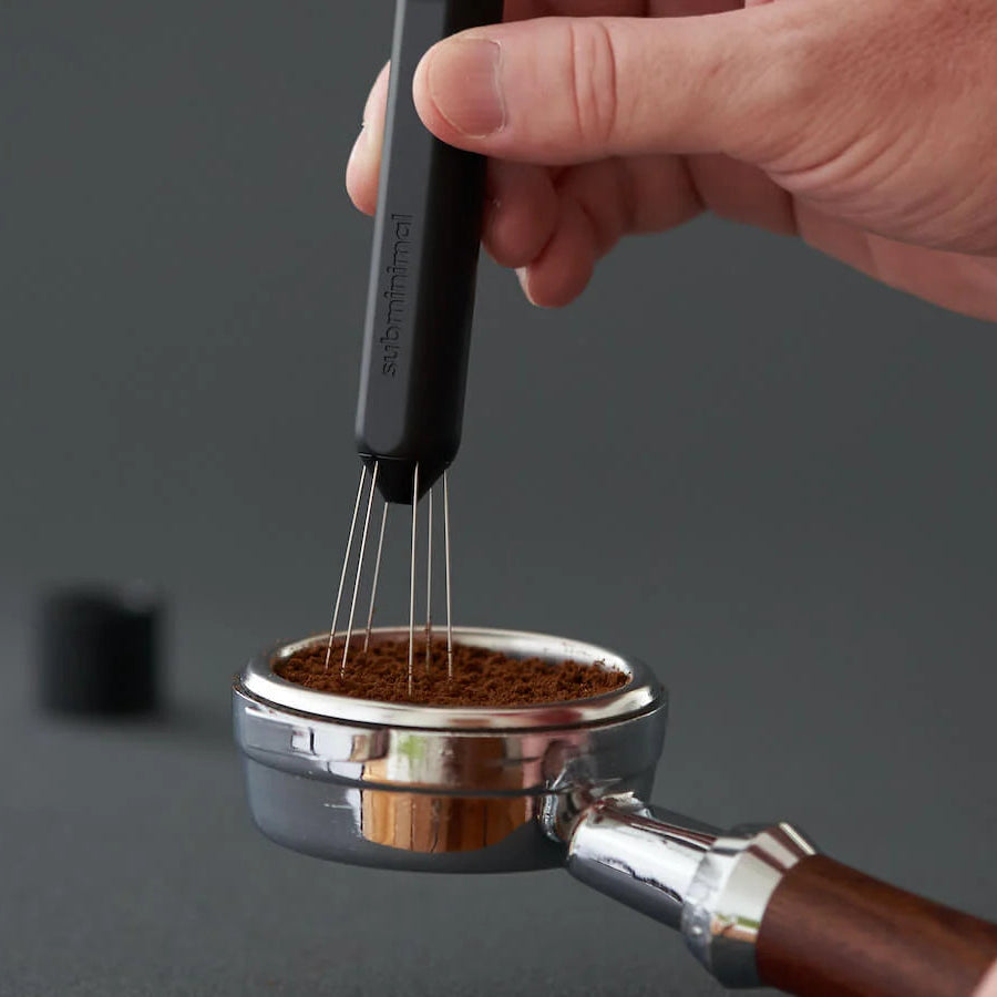 A hand holds a black coffee whisk over a stainless steel portafilter filled with ground coffee, with a dark gray background.