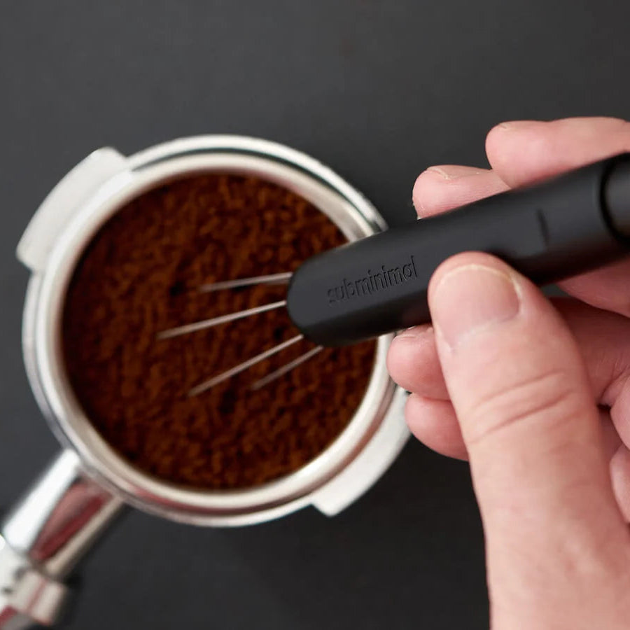 A hand holding a black 3-in-1 espresso whisk with the brand name 'espressomini' printed on the handle, positioned over a metal portafilter filled with ground coffee on a dark background.