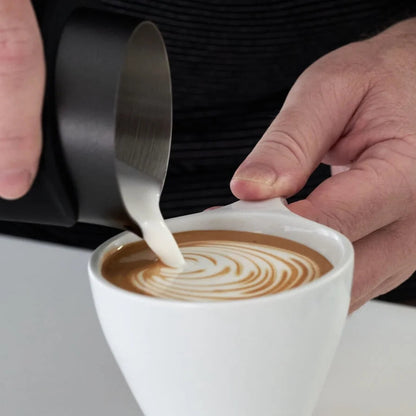 A close-up of a person pouring milk from a stainless steel pitcher into a white cup of coffee with latte art, set against a dark shirt and light tabletop background.