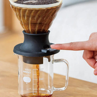 A hand operating a manual pour-over coffee maker with a conical paper filter, dark coffee grounds, a black plastic body, and a clear glass measuring cup on a wooden table.